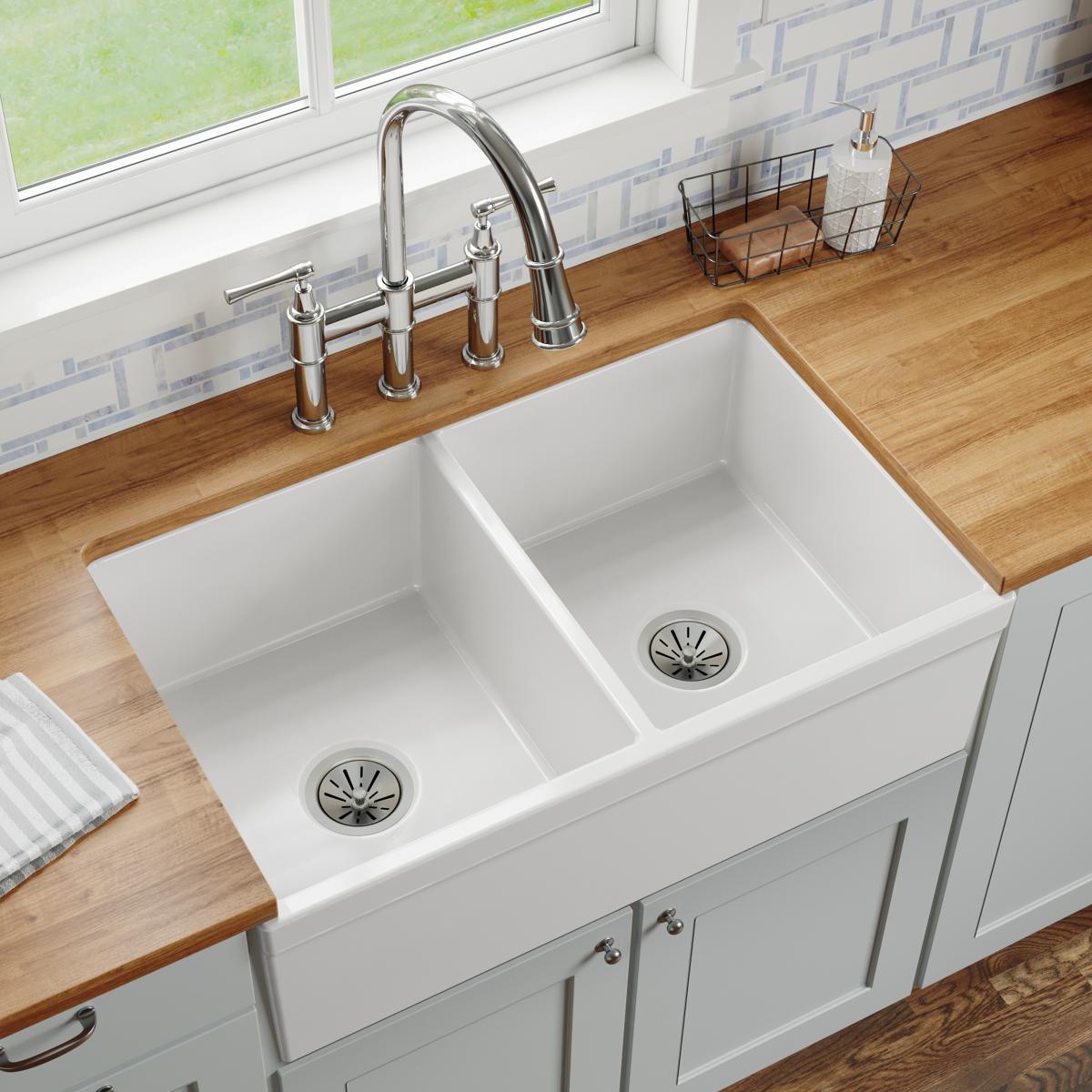 White fireclay farmhouse sink with two equal bowls and drain strainers, installed with chrome faucet on wood countertop.