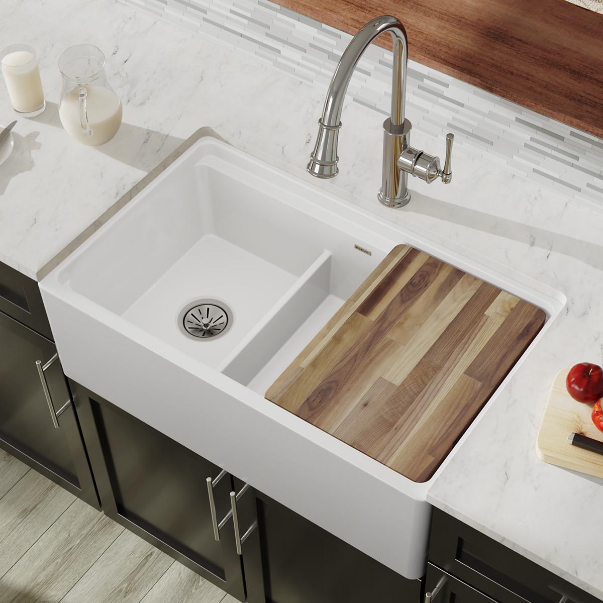 White fireclay farmhouse sink with two unequal bowls, one with drain and one with wood cutting board insert, paired with stai