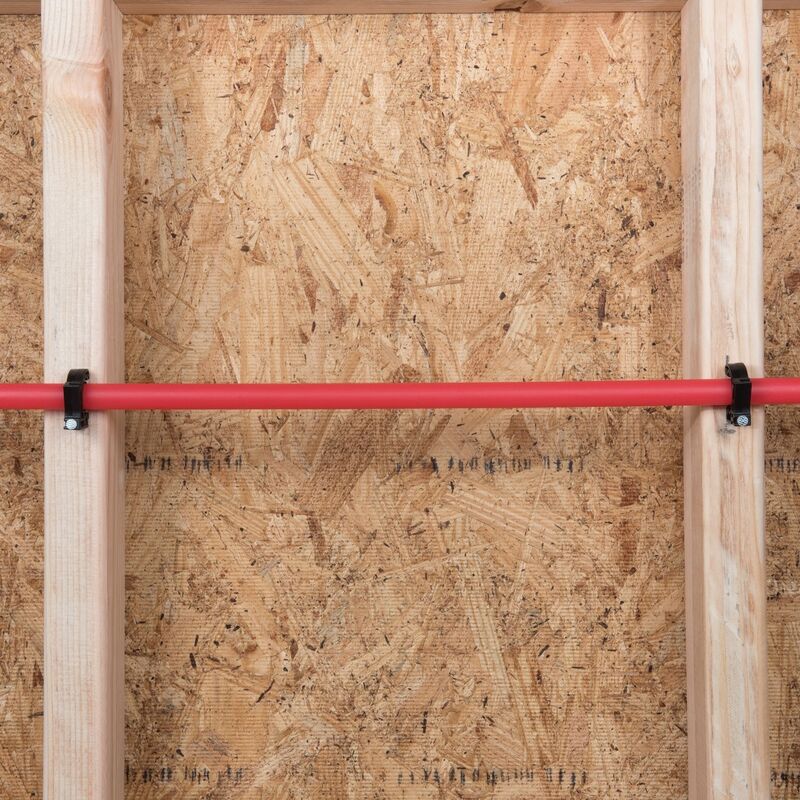 Steel half clamp with barbed nail securing a red pipe to wooden framing.