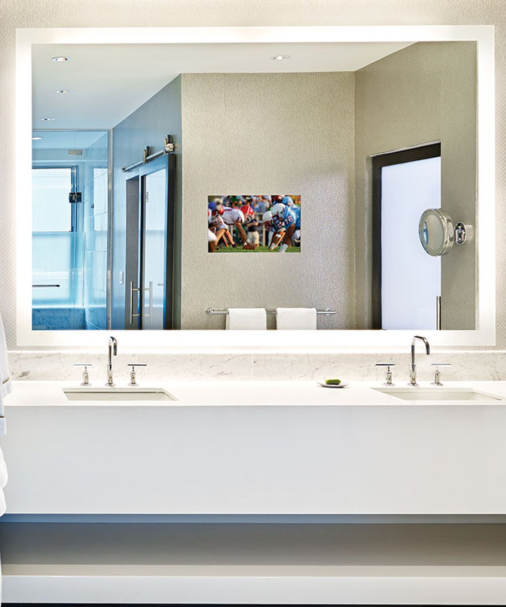 Large rectangular bathroom mirror with integrated TV display showing sports content, mounted above double sink vanity.