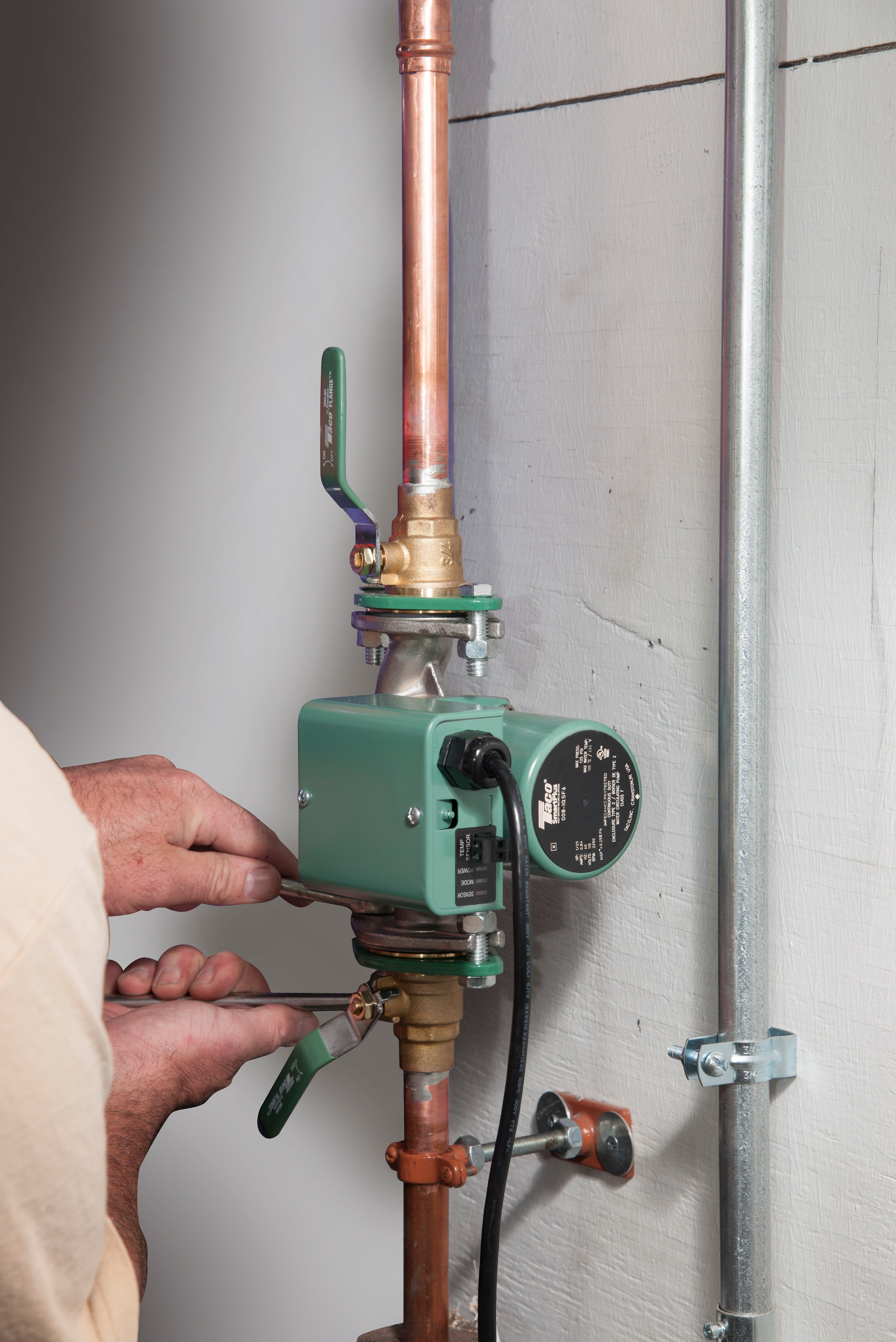 Green Taco hot water recirculating pump with integral check valve installed on copper piping with hand holding valve lever.