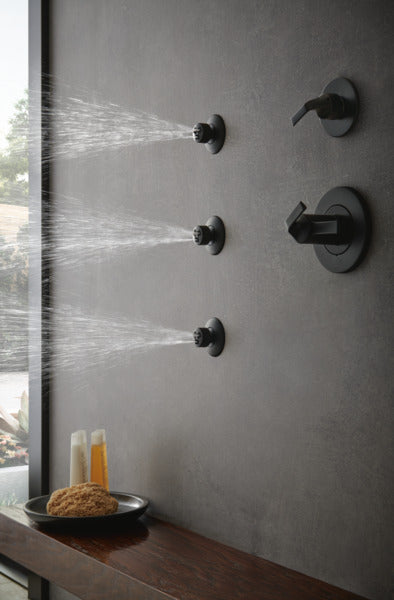 Matte black shower body jets mounted on gray tile wall with water spray visible from three nozzles.