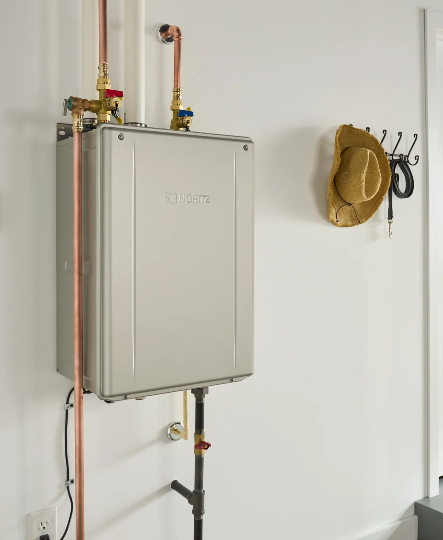 Tankless water heater mounted on a wall with visible pipes and a hat hanging nearby.