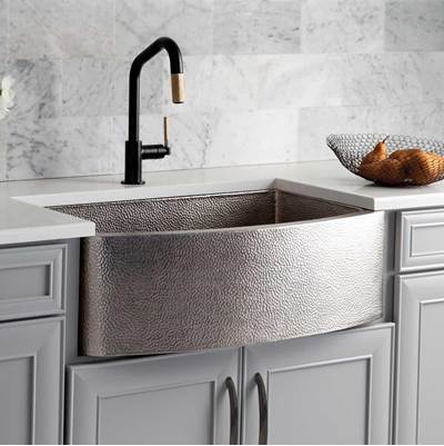 Brushed nickel farmhouse sink with curved front, black pull-down faucet, installed in white cabinetry with marble backsplash.