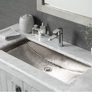 Polished nickel trough sink with hammered surface texture mounted in white marble countertop with faucet and drain.