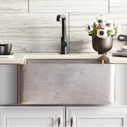 Brushed nickel farmhouse sink with hammered copper exterior and single black faucet installed in white cabinetry.