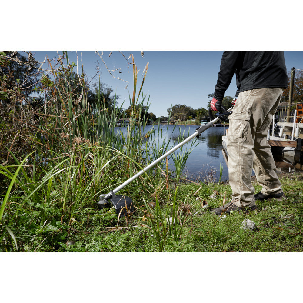 Person using brush cutter with blade attachment to trim tall grass and vegetation near waterside.