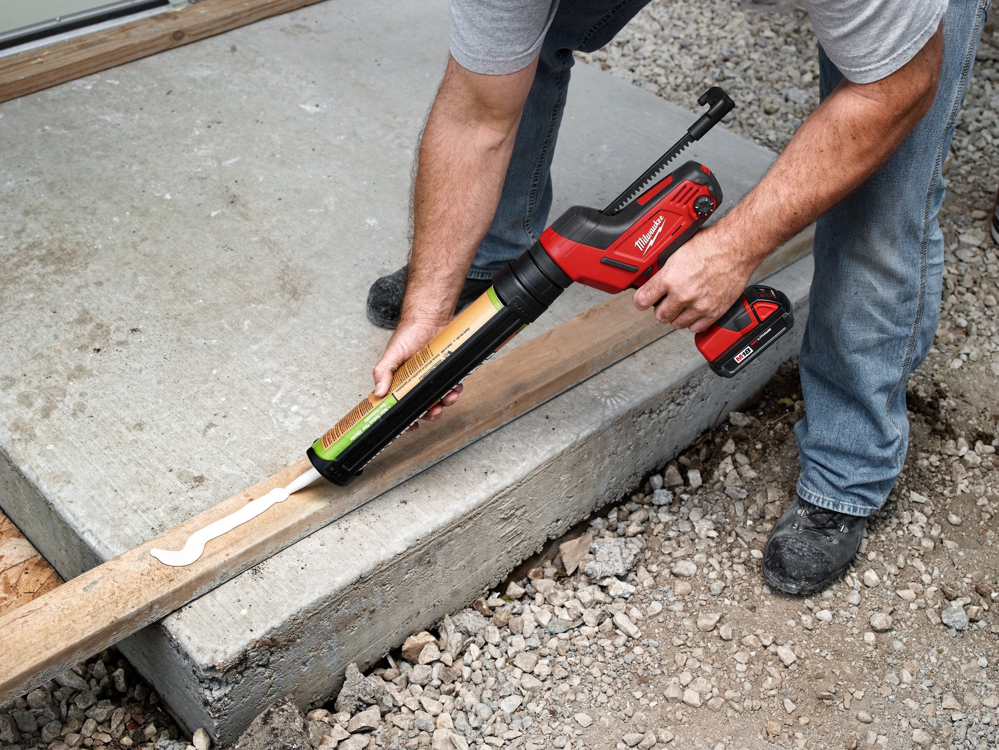 Red and black caulking gun with yellow and black aluminum barrel being used to apply sealant along a concrete edge.