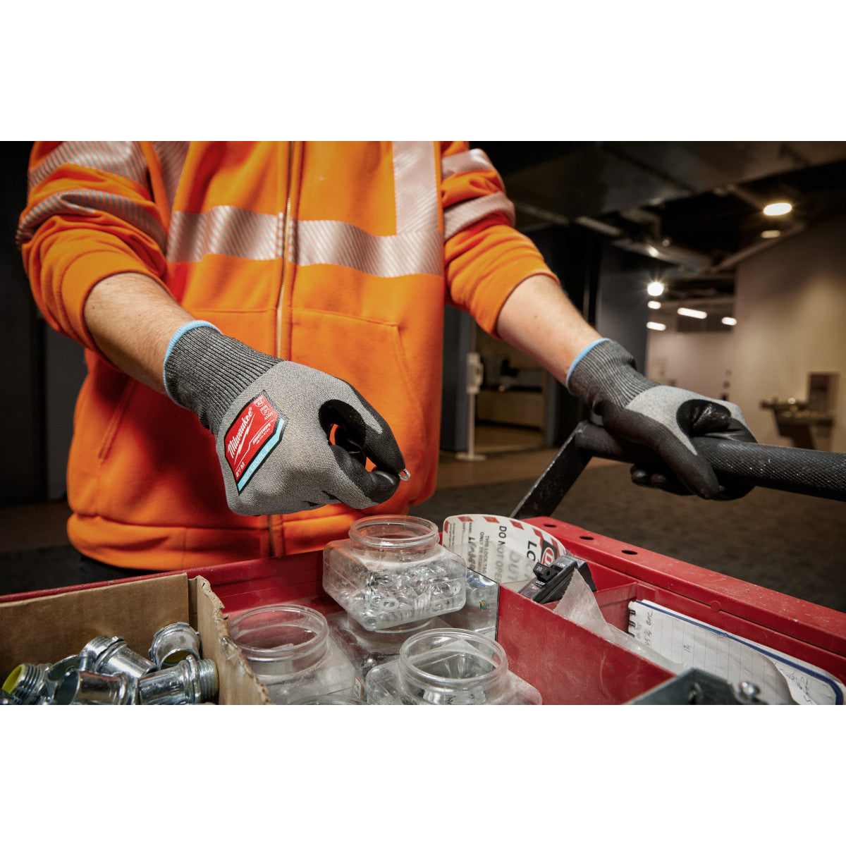 Worker wearing gray and black polyurethane dipped gloves with red Milwaukee branding while handling small parts in a toolbox.