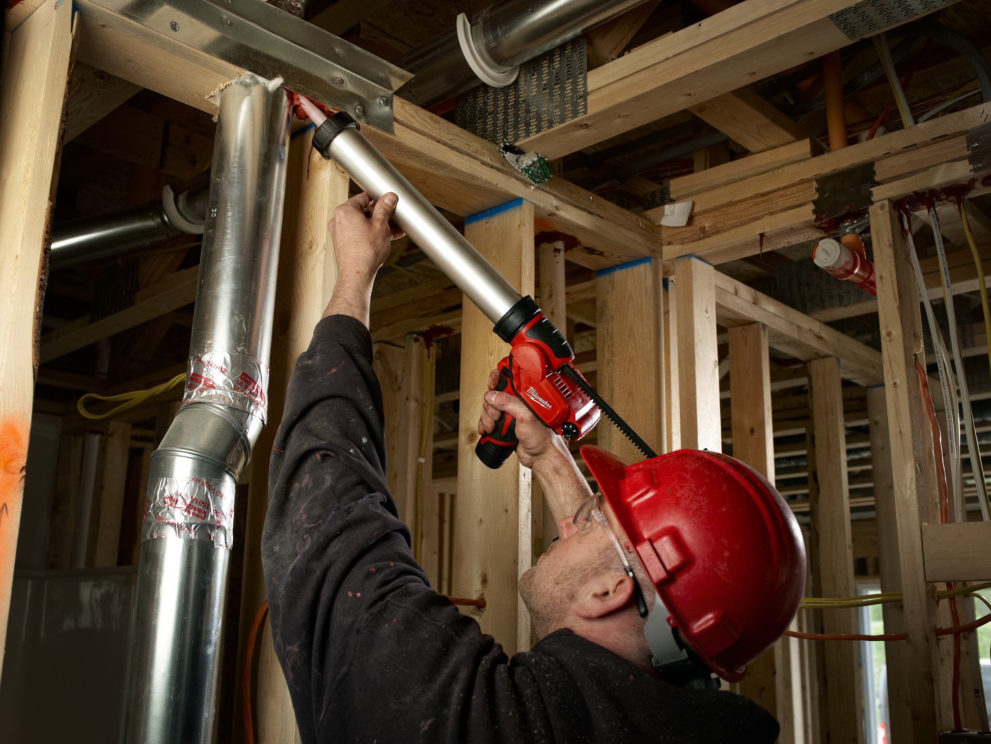 Worker in red hard hat using red caulking gun to apply sealant to aluminum barrel mounted on wooden frame.