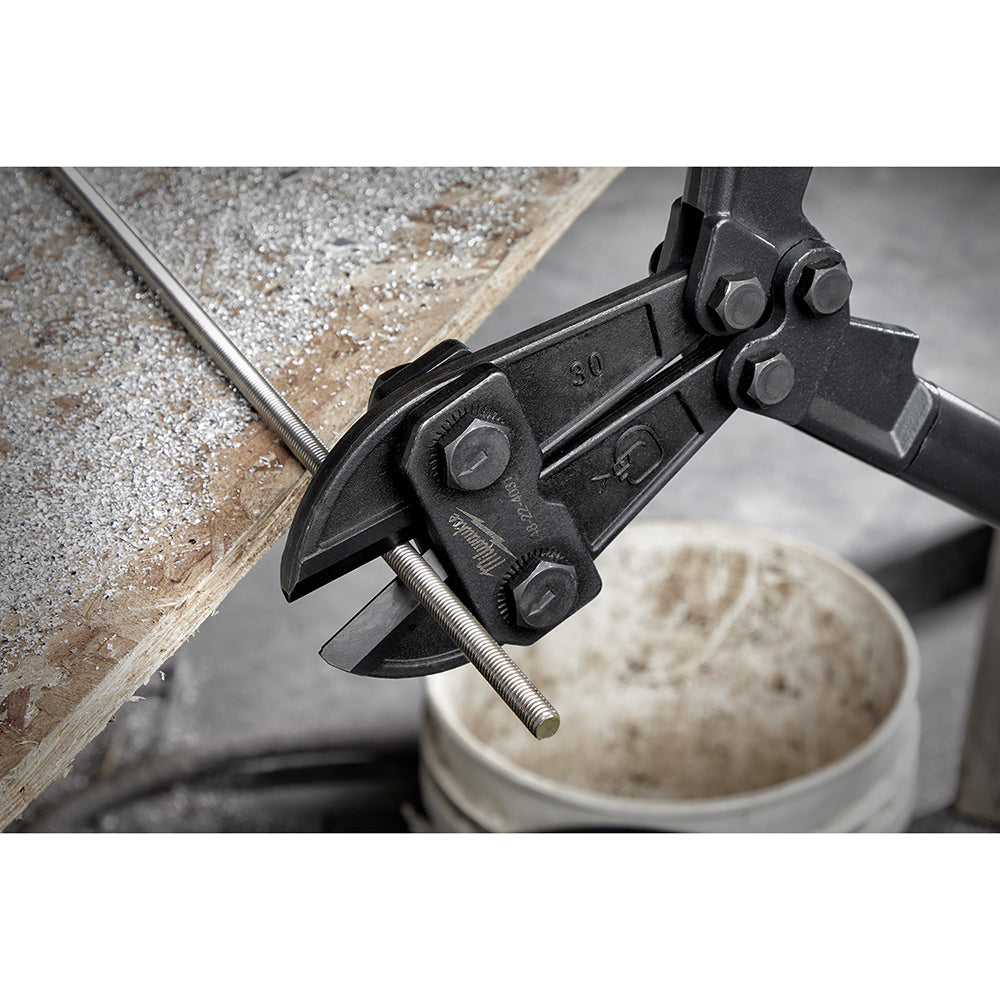 Black bolt cutter with metal handles cutting through a threaded bolt on a wooden surface.