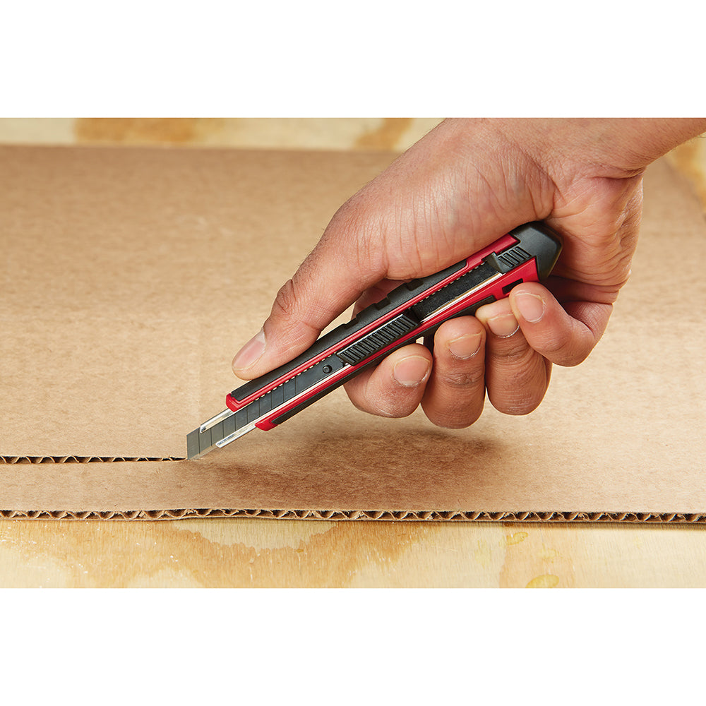 Red and black snap-off utility knife with metal blade cutting through cardboard box.