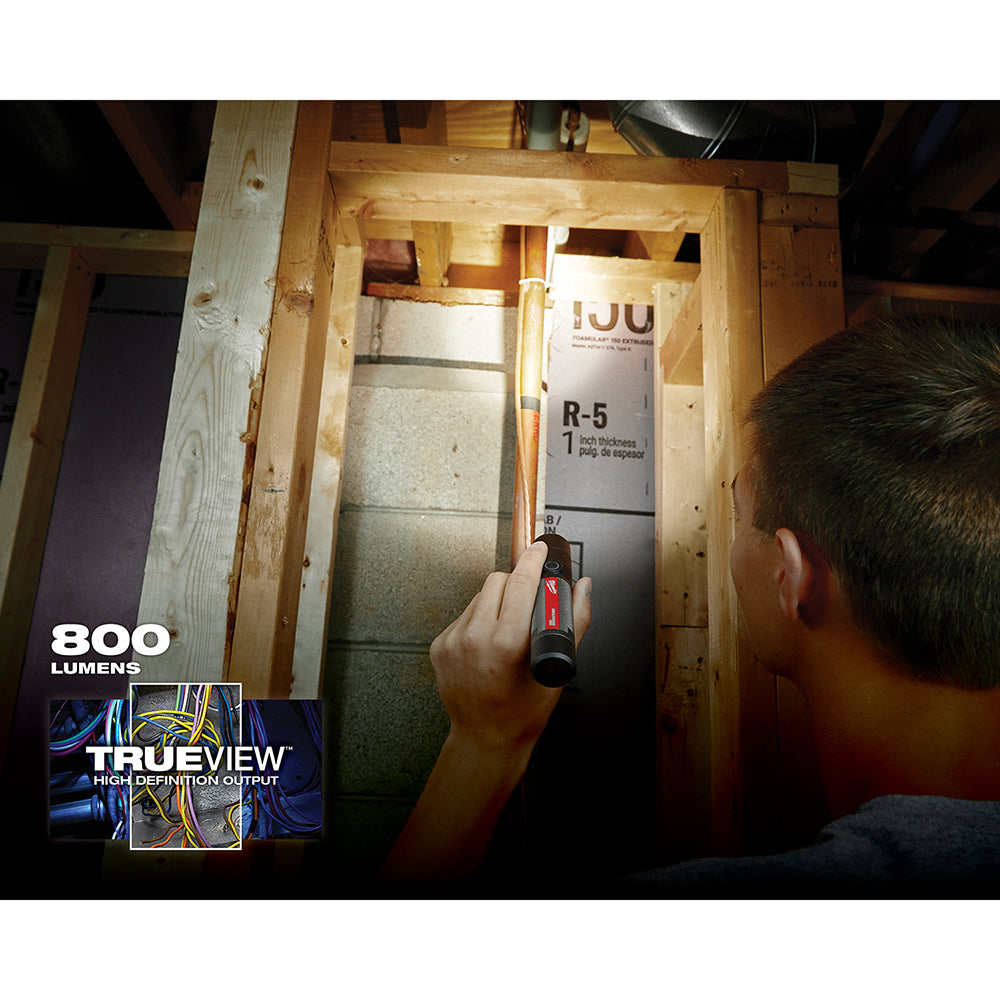 Hand holding compact Milwaukee flashlight illuminating insulation in wooden wall cavity.