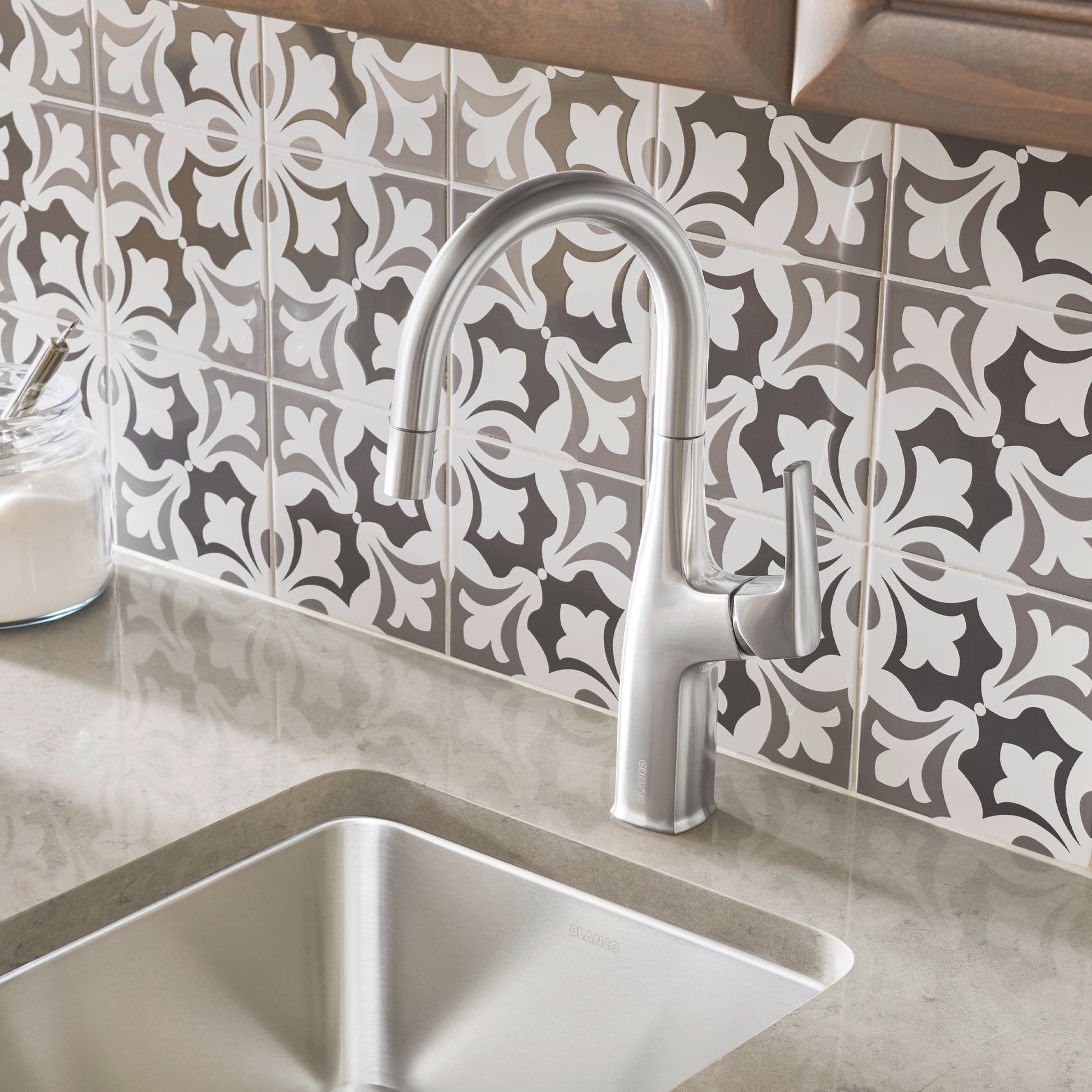 Stainless steel bar faucet with curved spout mounted above undermount sink on patterned tile backsplash.