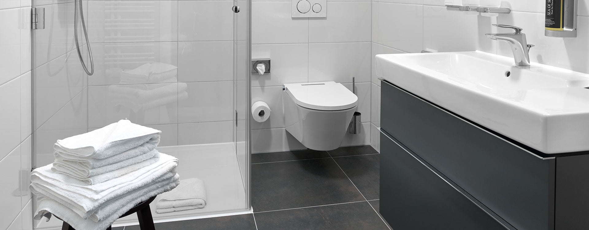 White wall-hung toilet with chrome fixtures in modern bathroom with dark tile flooring.