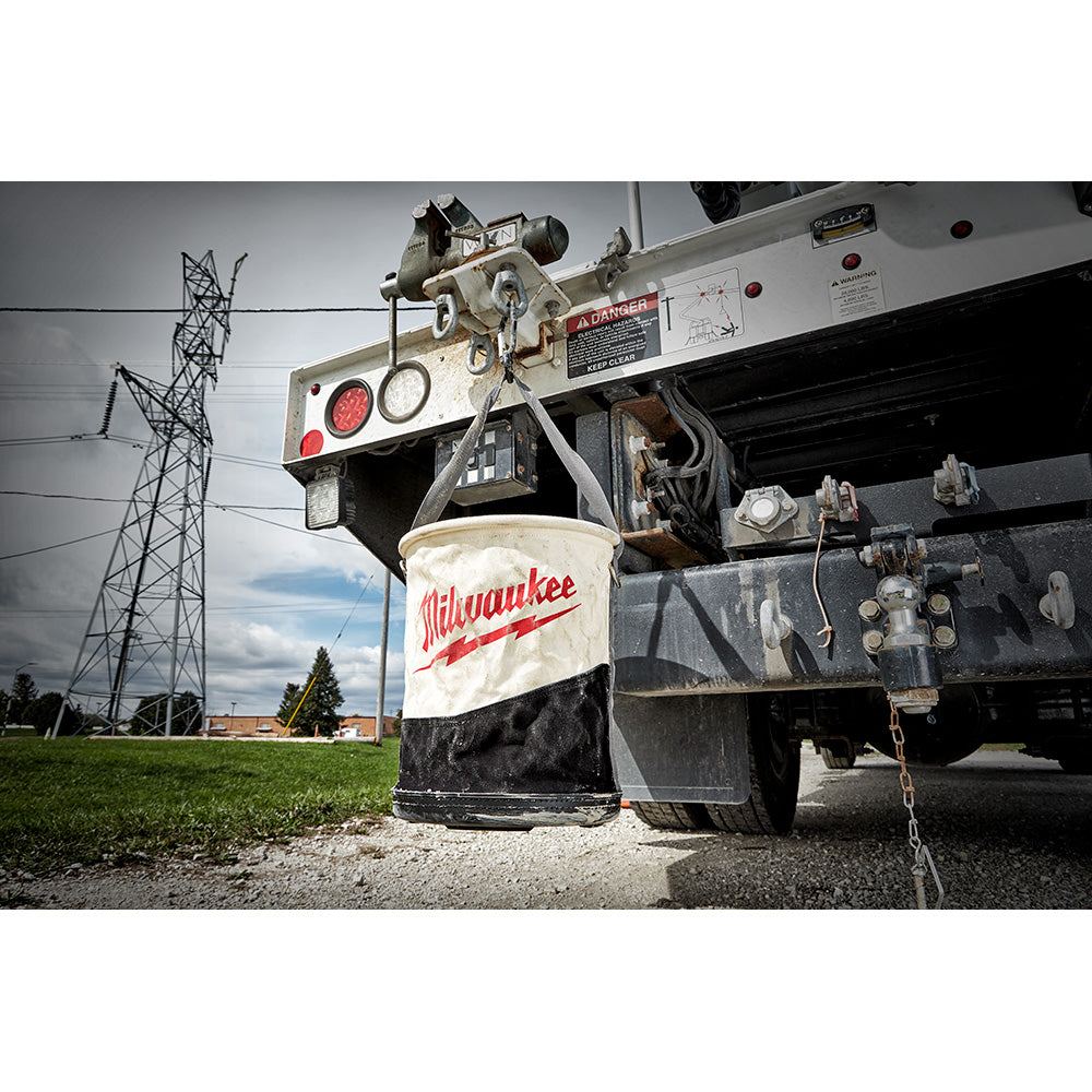 Canvas utility bucket with Milwaukee branding in cream and black, suspended from industrial lifting equipment.