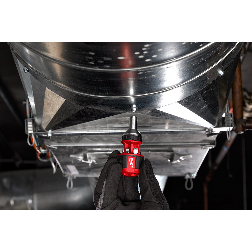 Red and black ratcheting multi-bit driver held in gloved hand against large metal industrial equipment.