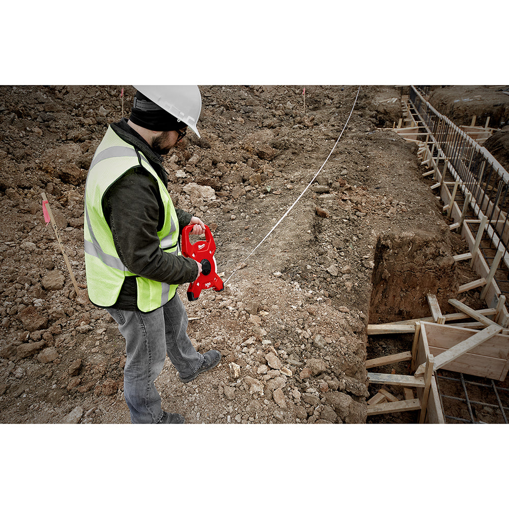 Worker in safety gear holding red open reel fiberglass tape measure on construction site.