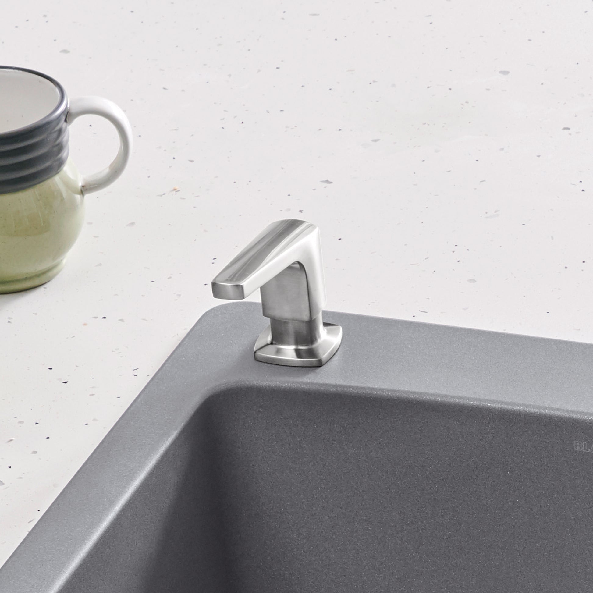 Stainless steel soap dispenser with angular pump mounted on gray sink rim next to ceramic mug.