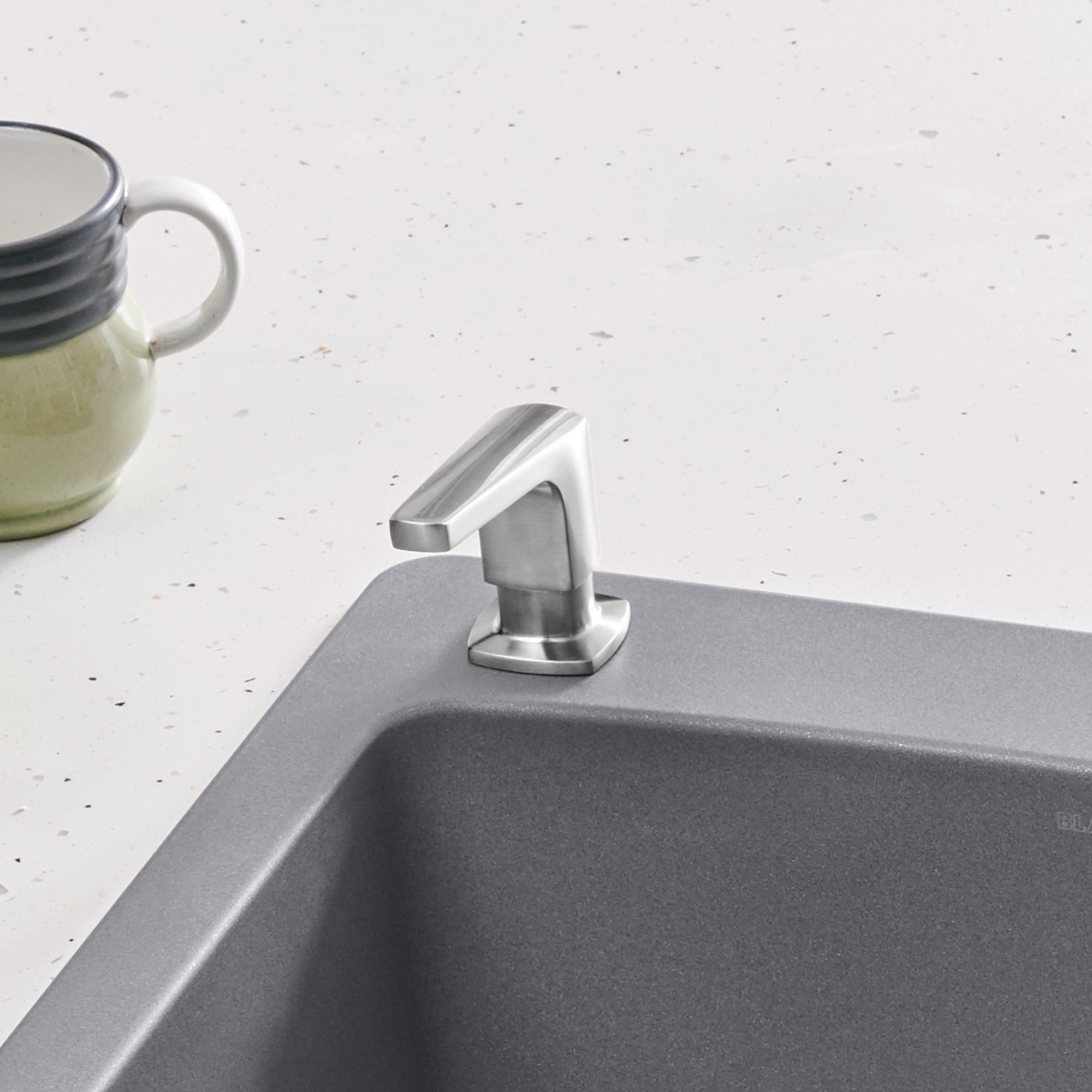 Stainless steel soap dispenser with angular pump mounted on gray sink rim next to ceramic mug.