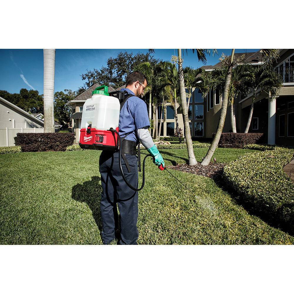 Person wearing backpack sprayer with attached hose and spray wand extended toward ground in residential yard.