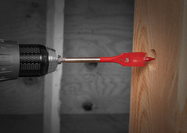 Red spade bit with metal shaft inserted in drill, drilling into wood surface.