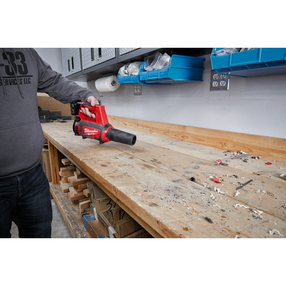 Milwaukee M12 compact spot blower with red housing and black nozzle, held in hand above wooden workbench.