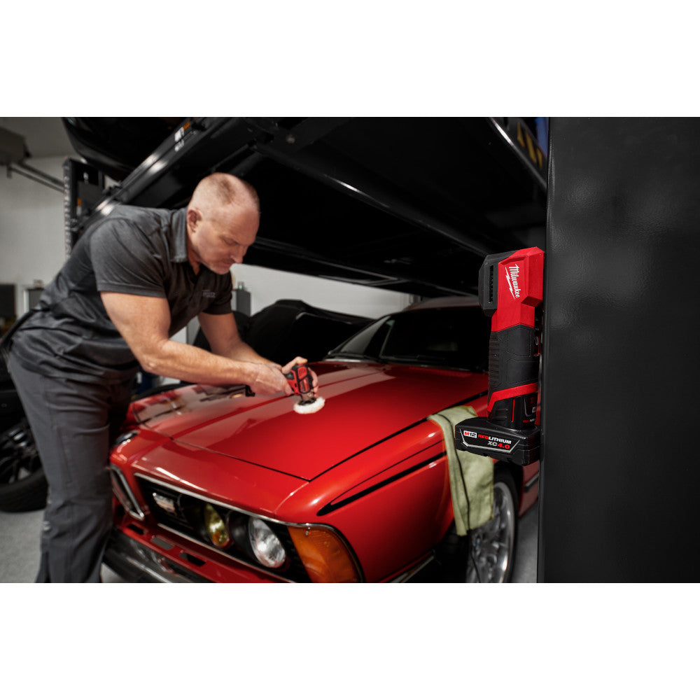 Milwaukee M12 paint and detailing color match light mounted on garage wall while technician inspects red vehicle hood.