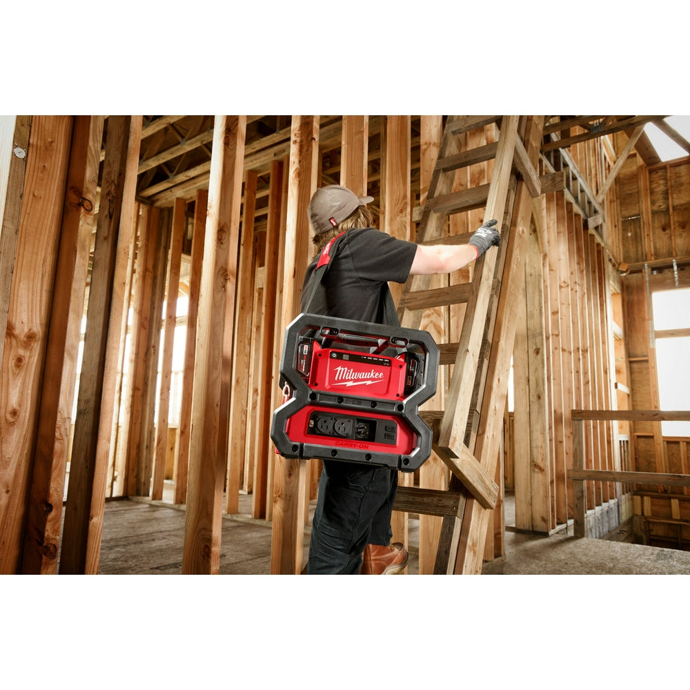 Worker wearing black shoulder strap across chest carrying red and black Milwaukee power supply unit on construction site.