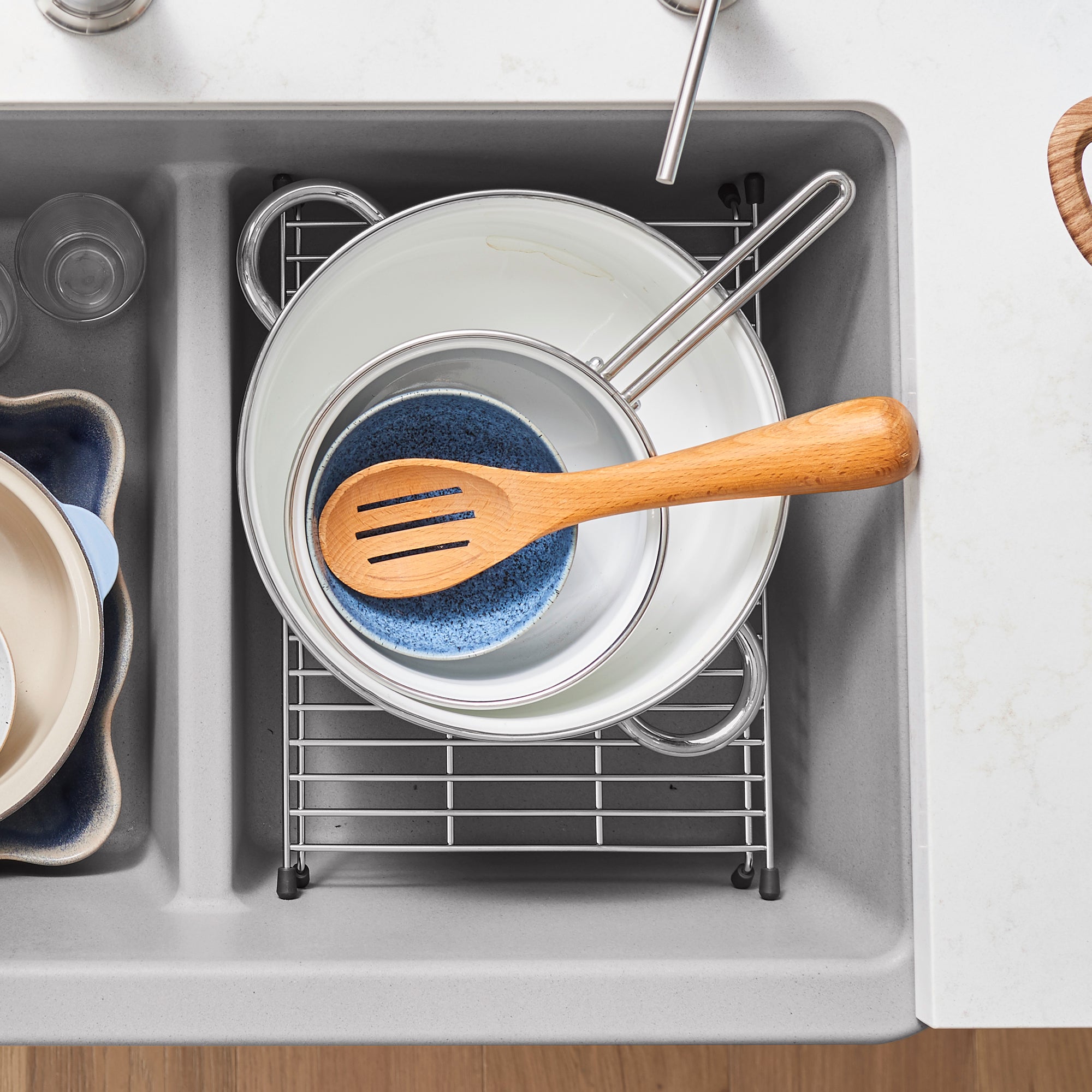 Stainless steel sink grid with white cookware and wooden utensil inside dark gray sink basin.