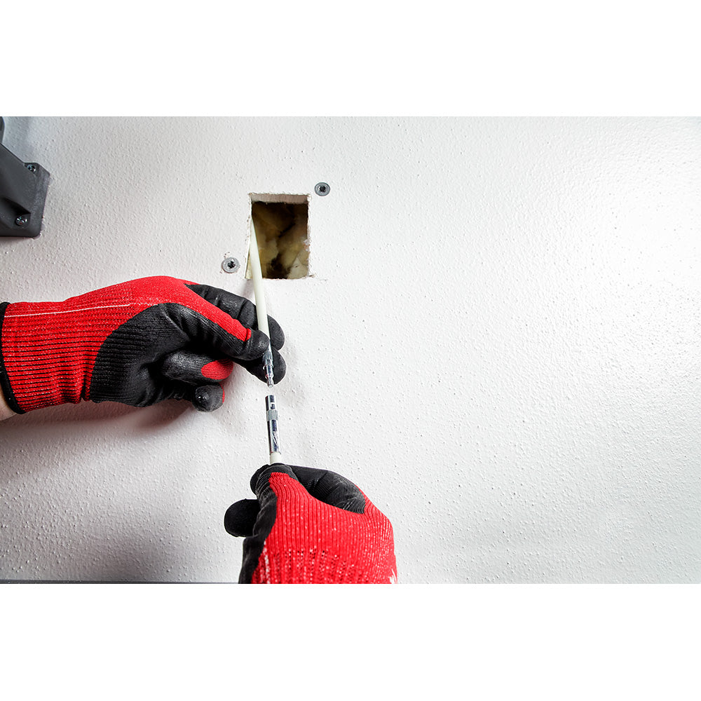 Hands in red and black gloves threading a white fish stick tool through a wall opening with electrical box visible.