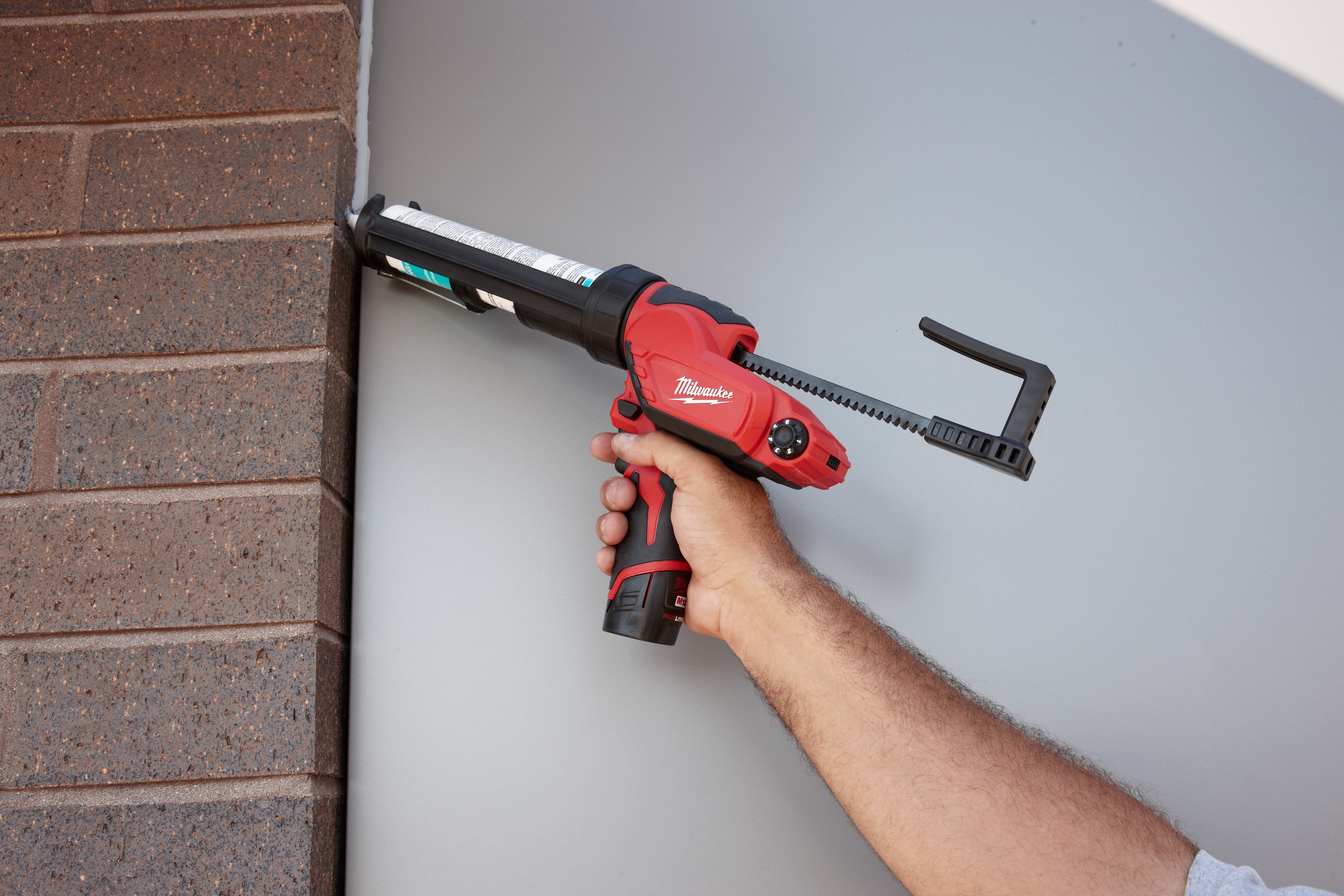 Red and black Milwaukee caulk gun with aluminum barrel held by hand against brick and white surface.