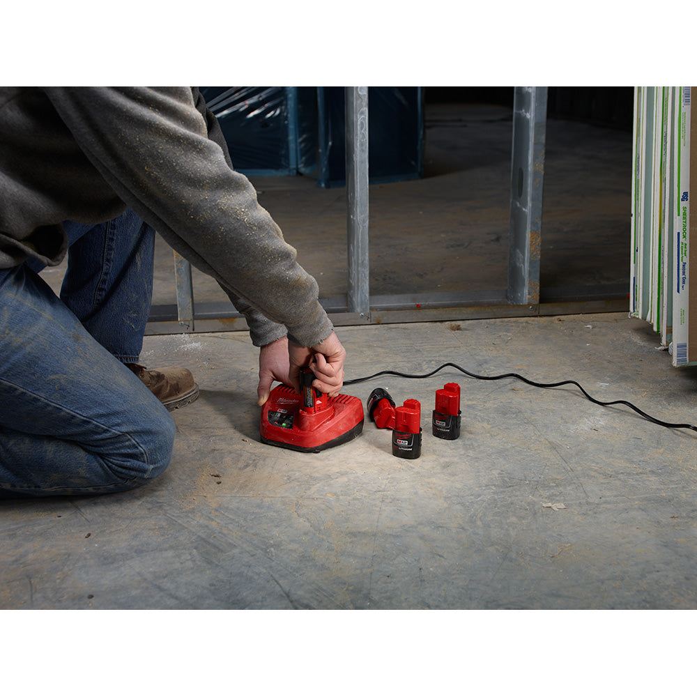 Red Milwaukee M12 battery charger with two black lithium-ion batteries positioned on concrete floor.
