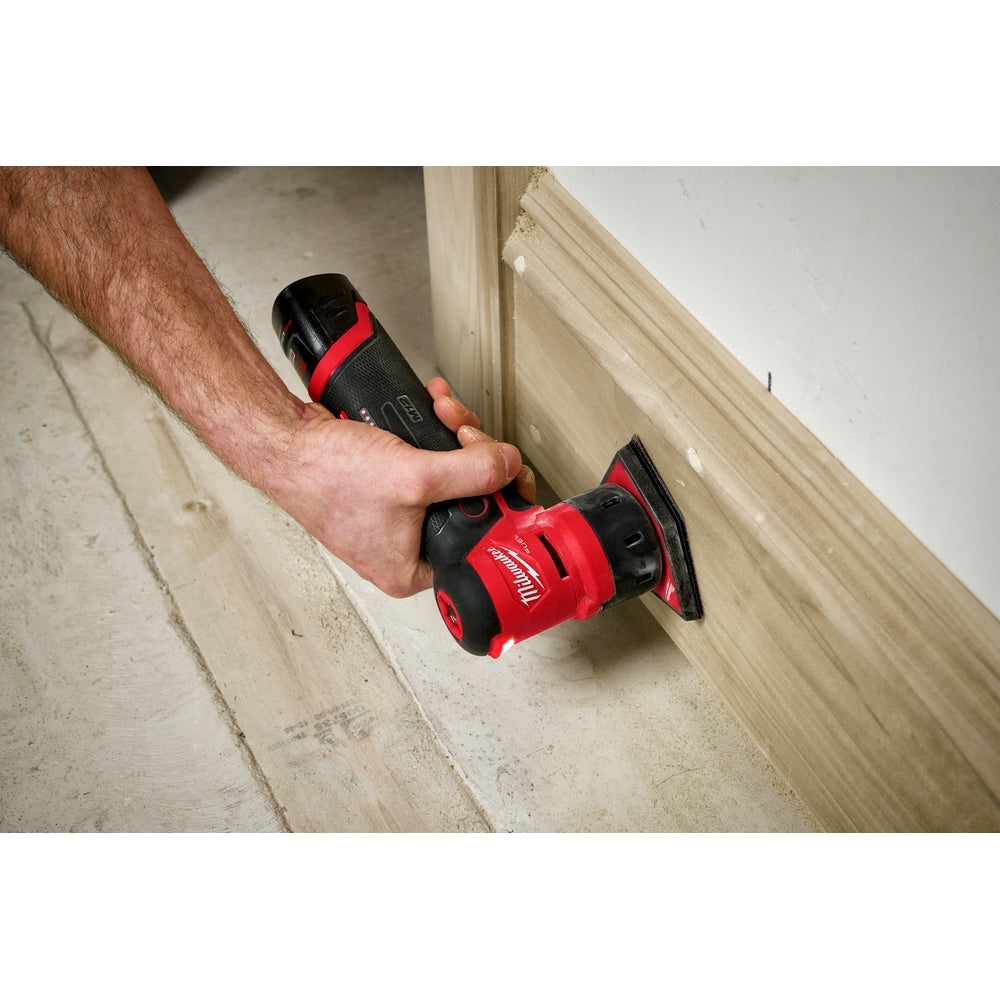 Red and black Milwaukee M12 orbital detail sander with rectangular pad being held against wooden trim.
