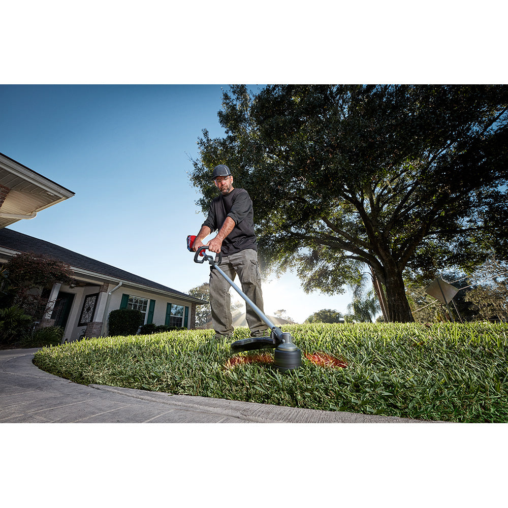 Man using red and black string trimmer on residential lawn with house and trees in background.