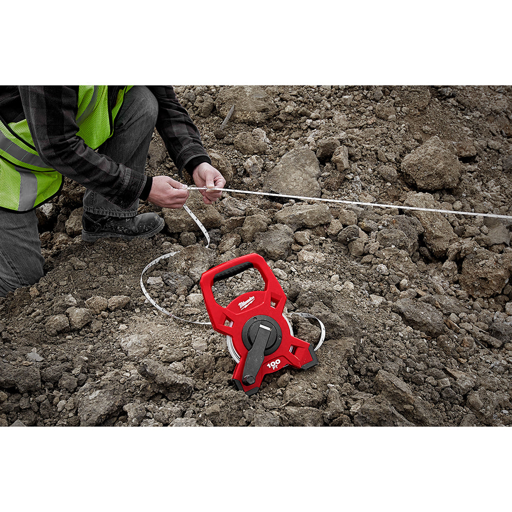 Red steel open reel tape measure with white measuring tape extended across rocky ground, held by worker in safety vest.