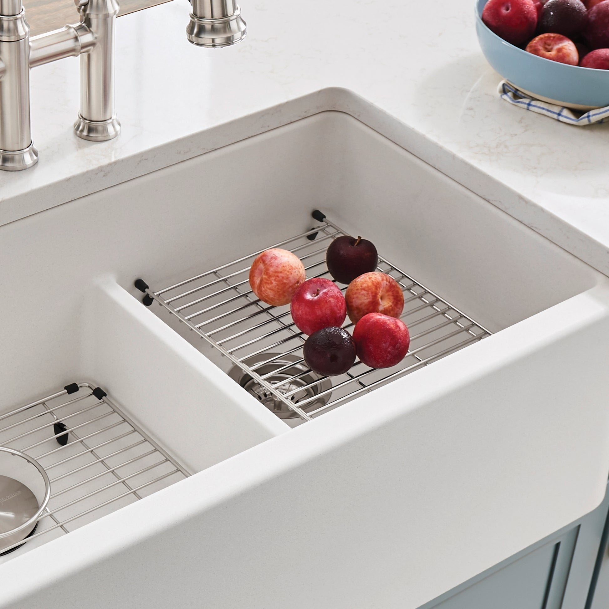 Stainless steel grid with black handles resting in white farmhouse sink basin, holding fresh apples.