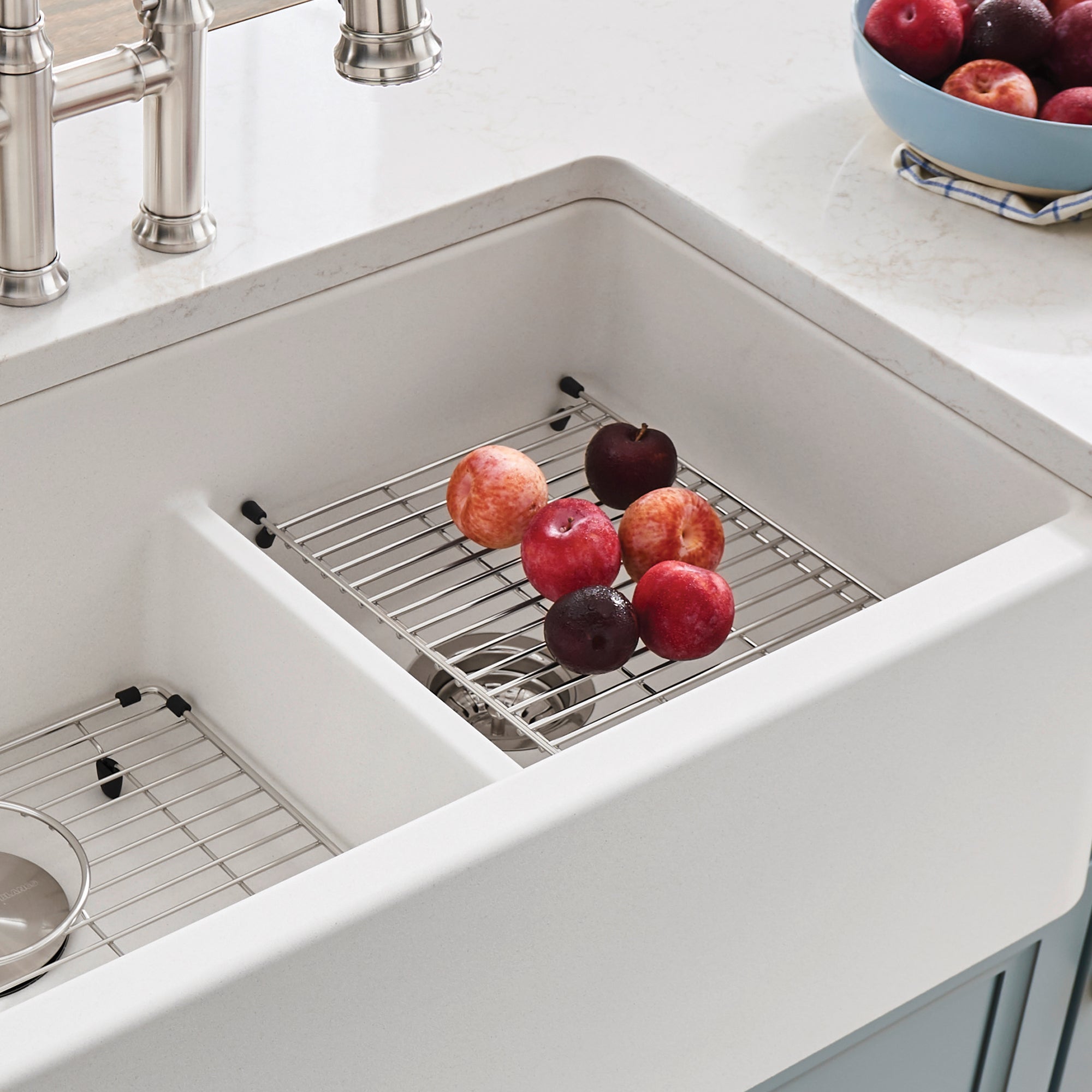 Stainless steel grid with black handles resting in white farmhouse sink basin, holding fresh apples.