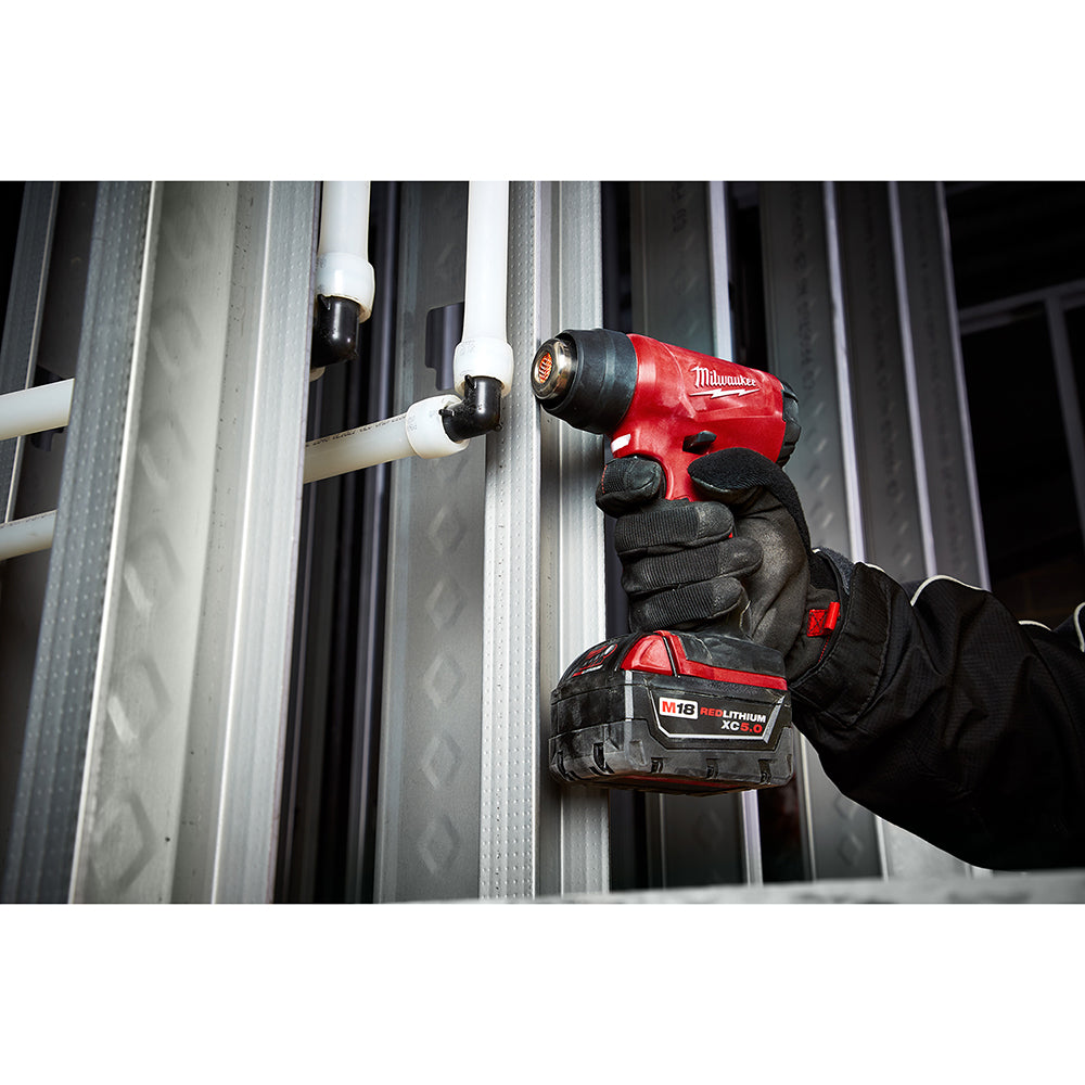 Red and black Milwaukee M18 compact heat gun held in gloved hand, directing heat toward white PVC pipe.