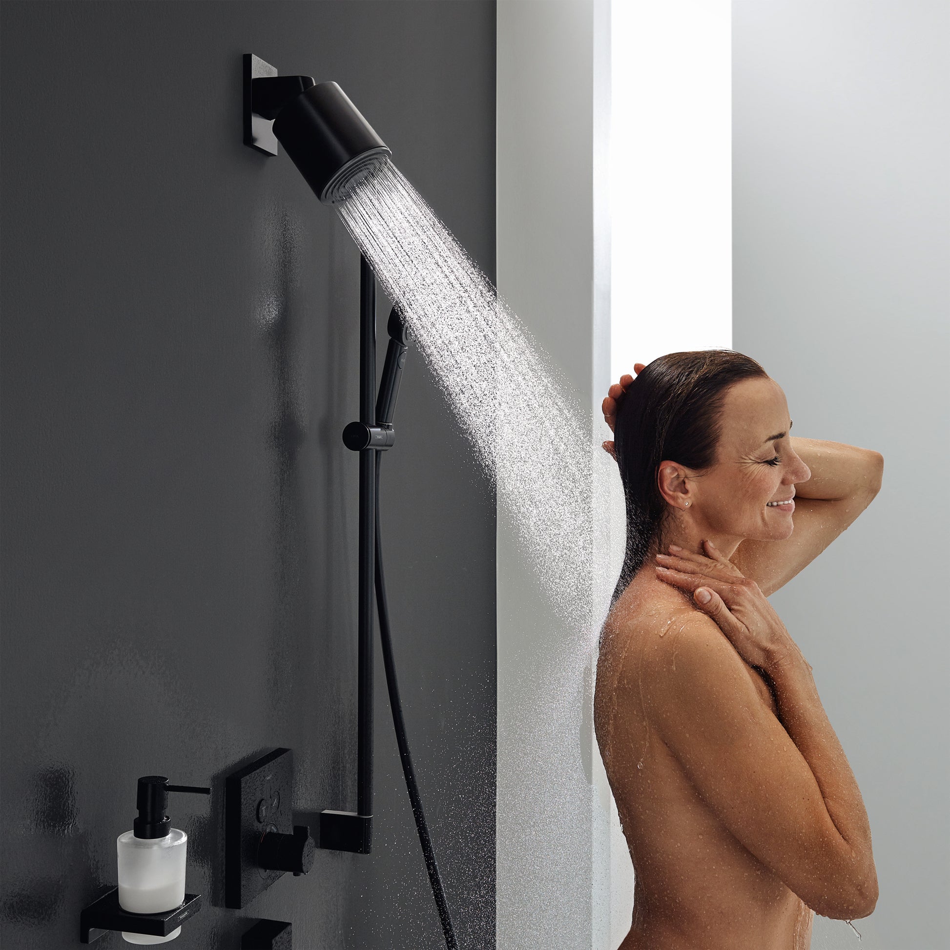 Matte black cylindrical shower head mounted on wall bracket with water stream flowing, accompanied by black wall-mounted cont