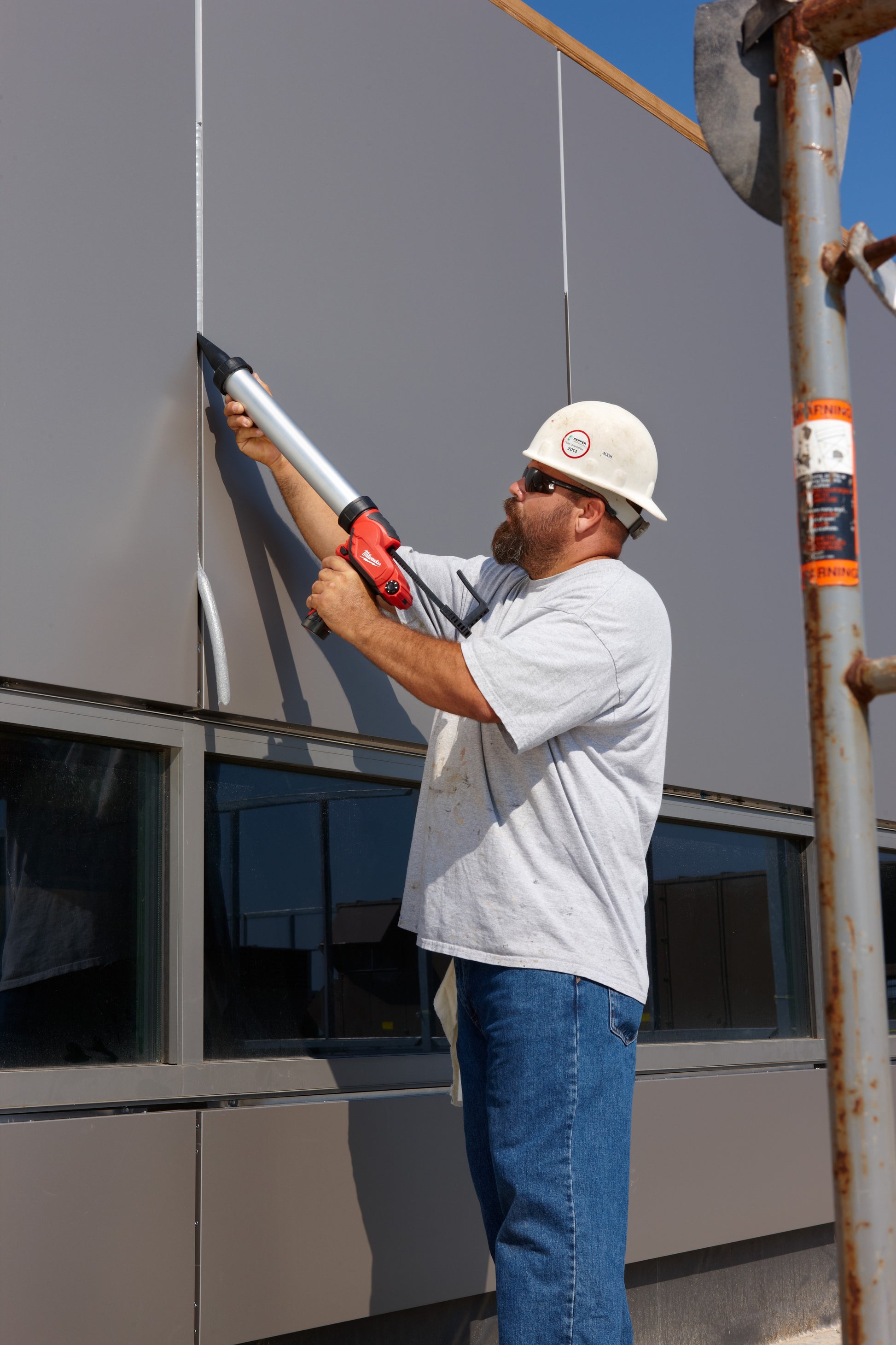 Worker holding aluminum barrel caulking gun with red handle against building exterior.