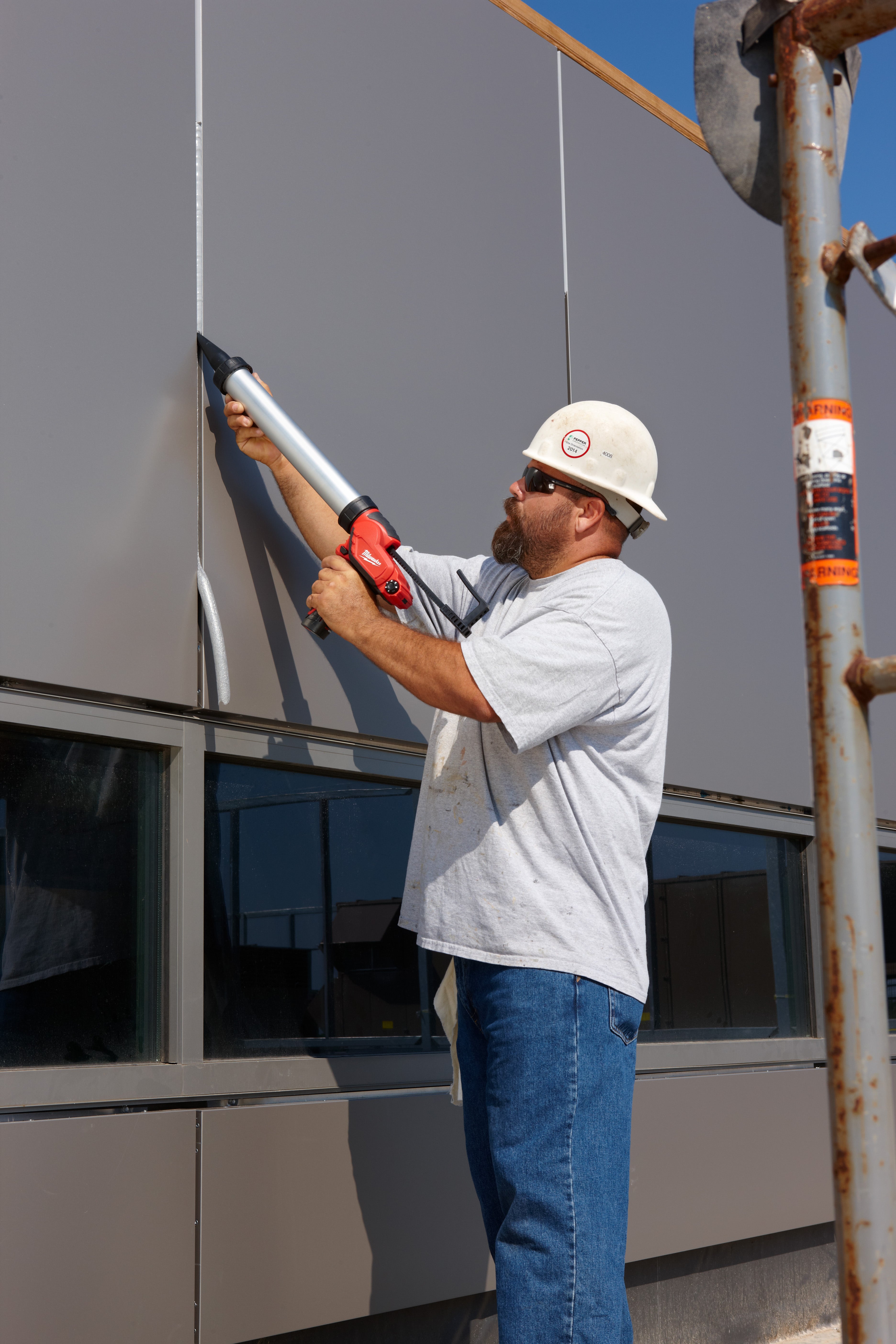 Worker holding aluminum barrel caulking gun with red handle against building exterior.