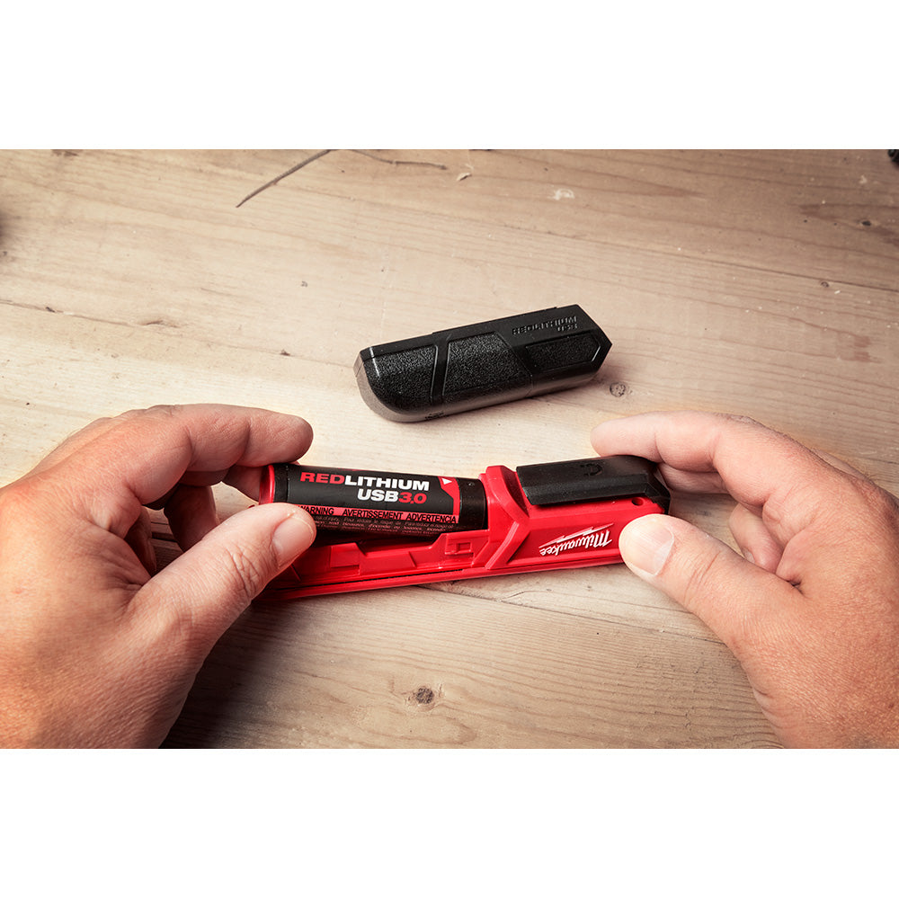 Two red and black Milwaukee REDLITHIUM USB batteries held in hands, with a third battery visible on wood surface.
