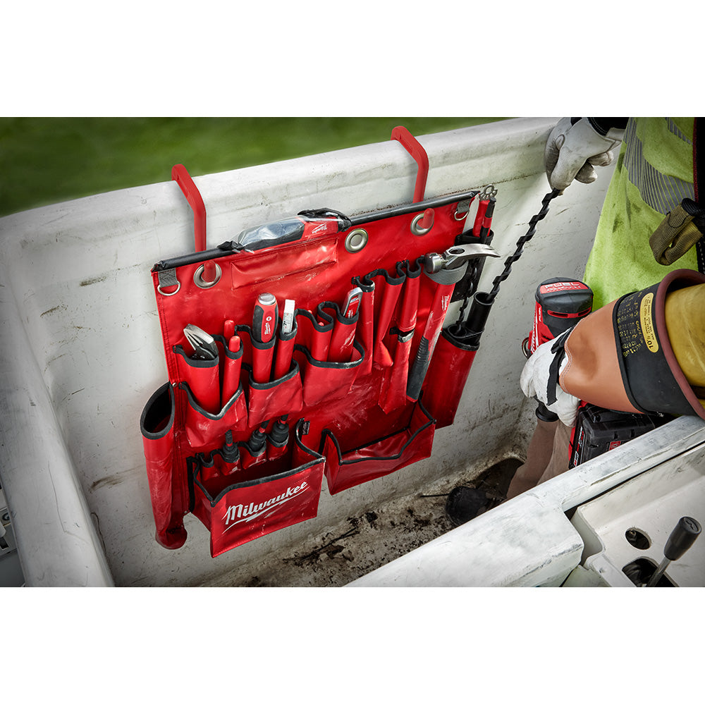 Red Milwaukee tool apron with multiple pockets and metal grommets hanging on white ladder rail.