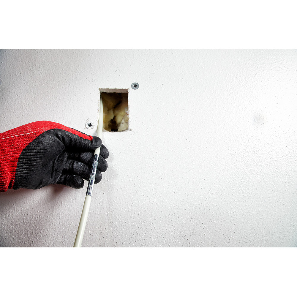Hand in black glove and red sleeve inserting white fish stick tool into wall opening with electrical box visible.