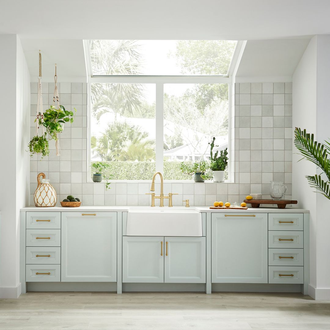 White apron front farmhouse sink with single gold faucet installed in light green kitchen cabinetry below subway tile backspl