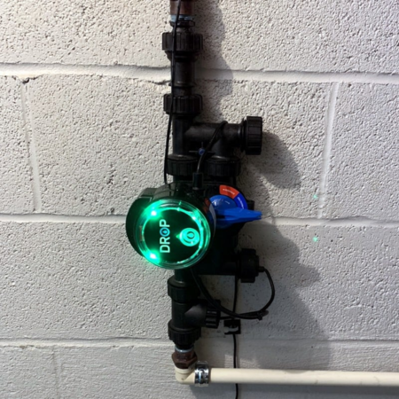 Black water shut-off valve with green illuminated display and red manual override lever mounted on white brick wall.