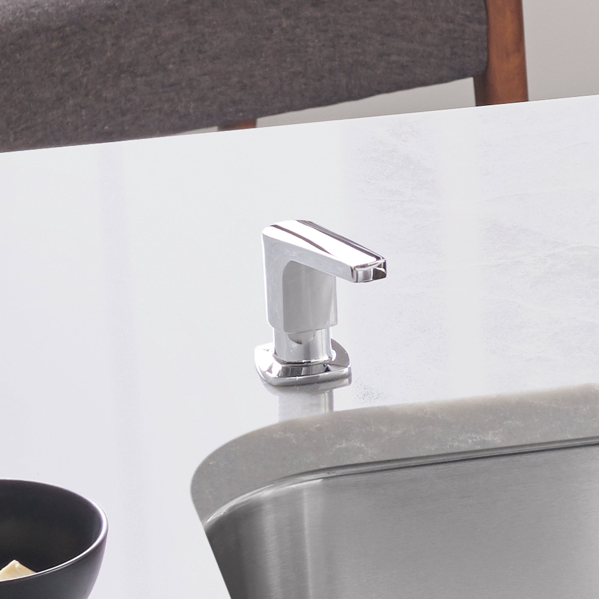 Chrome soap dispenser with white pump body mounted on bathroom counter near sink.