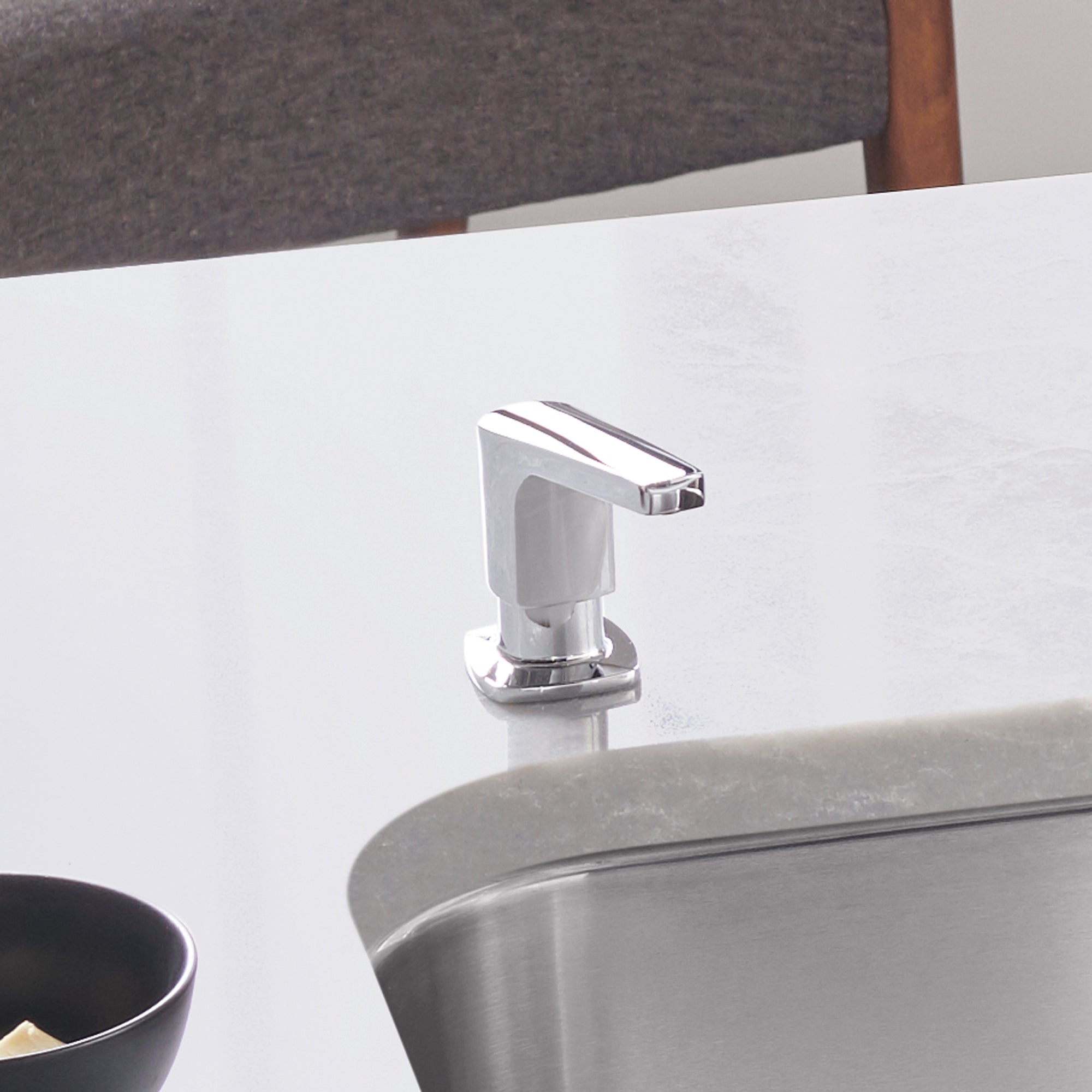 Chrome soap dispenser with white pump body mounted on bathroom counter near sink.