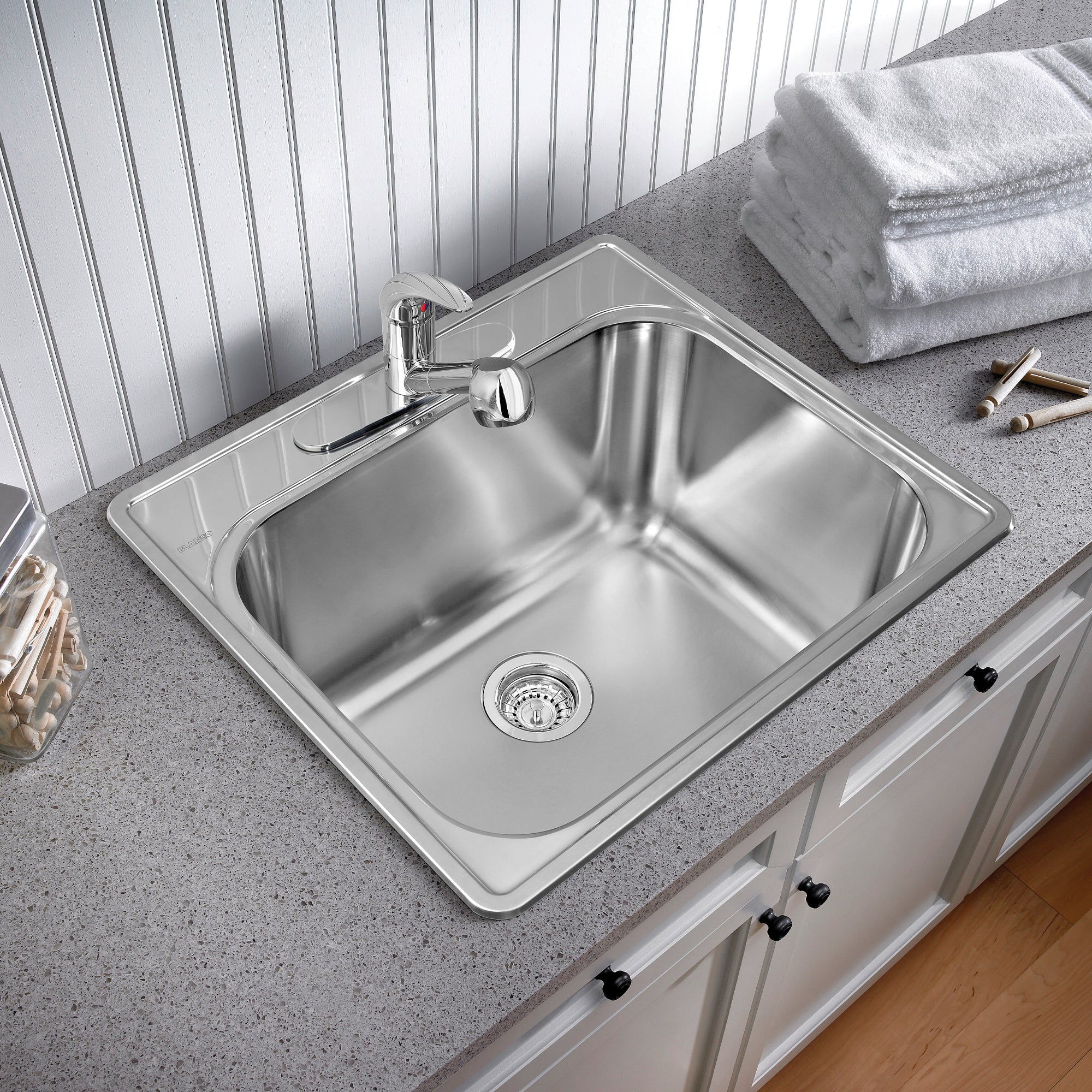 Stainless steel drop-in laundry sink with single basin, faucet, and drain strainer installed on gray countertop.